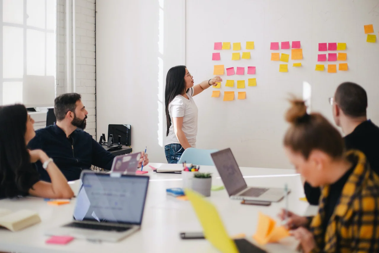 Workshop with a presenter arranging sticky notes on a whiteboard while four colleagues work at laptops