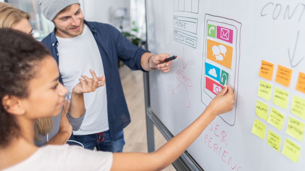 Three people at a whiteboard sketching a webpage wireframe with coloured sticky notes and marker drawings